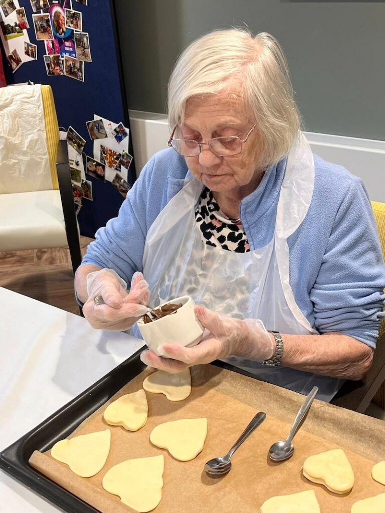 elderly lady wearing an apron and gloves decorating love heart biscuits with chocolate