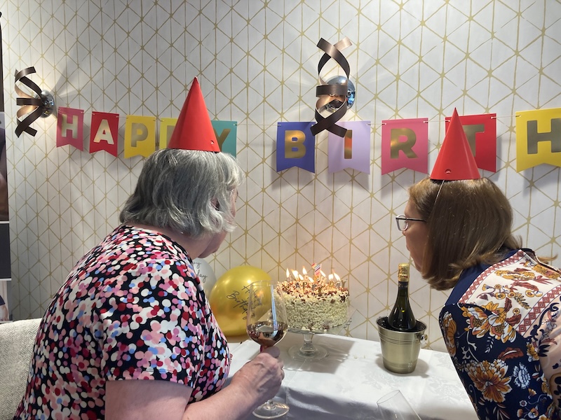 elderly resident and our home manager stood together in floral dresses blowing out candles on a cake