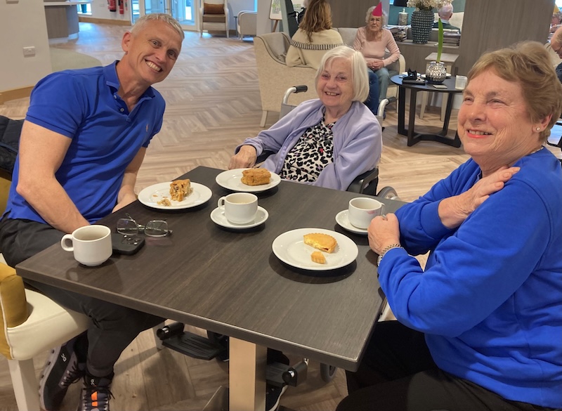 a female resident sat at a table with 2 of her relatives smiling with a slice of cake and a hot drink