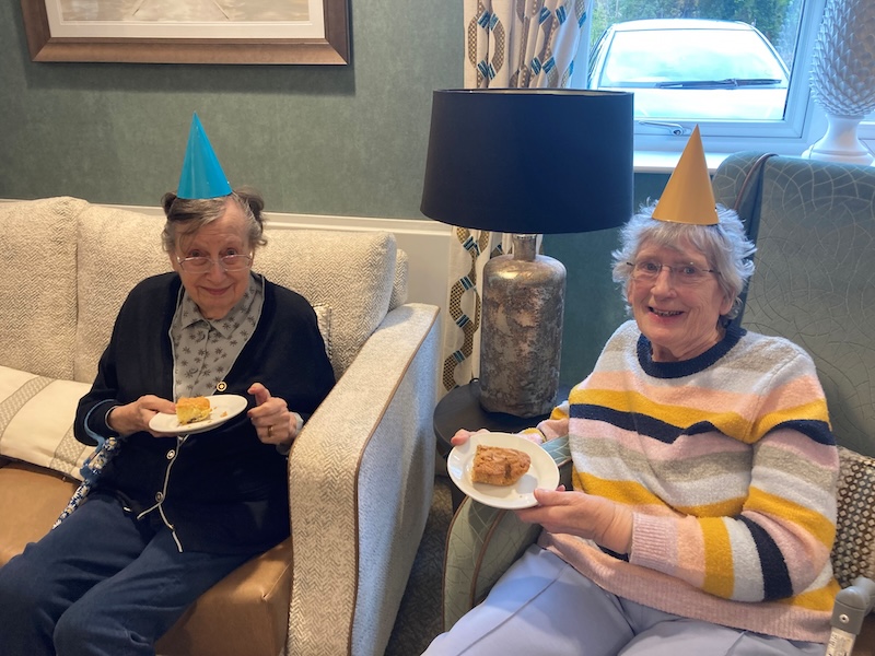2 of our female residents sat together in the lounge wearing party hats and smiling enjoying a slice of cake